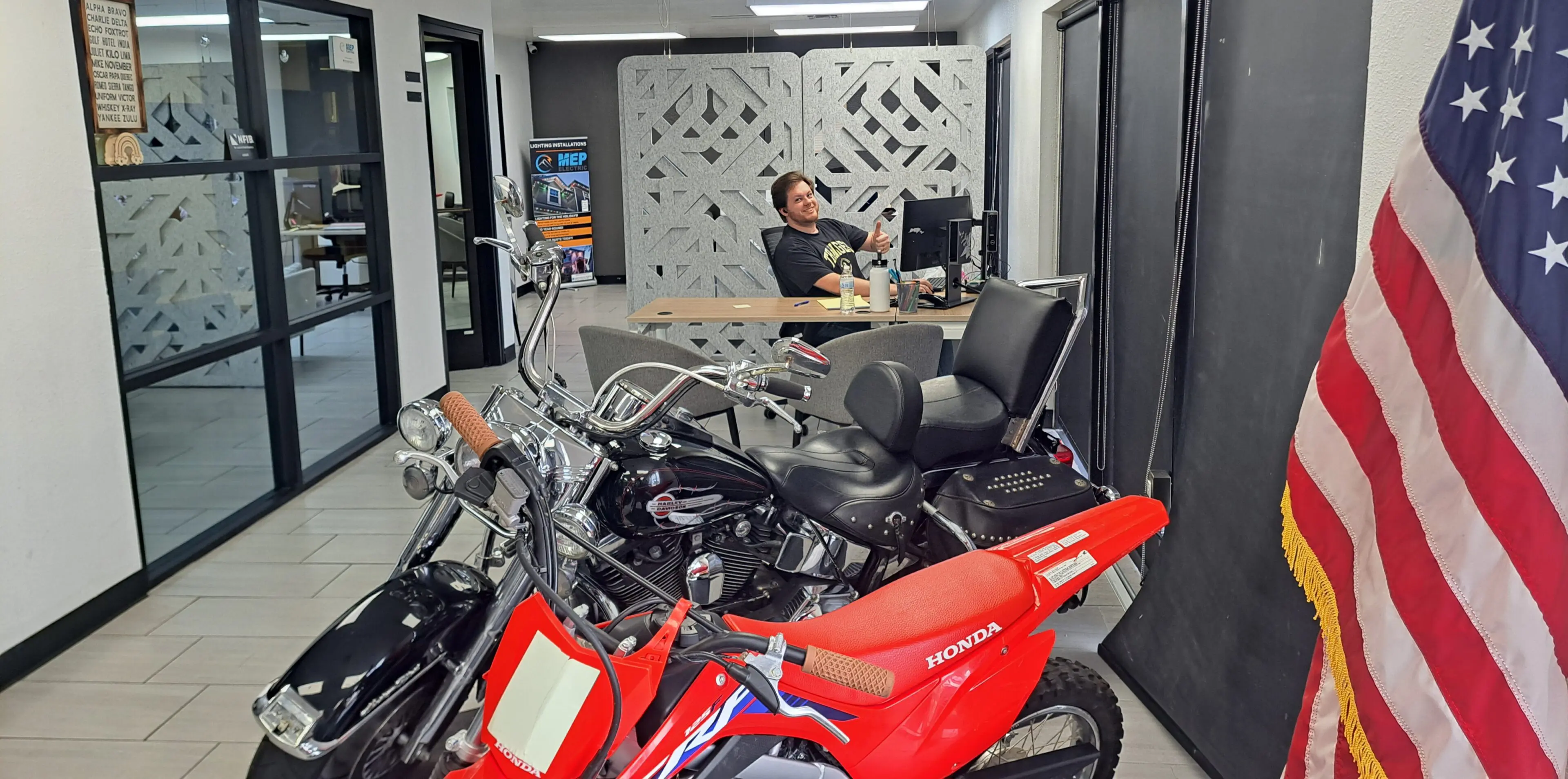 A large blonde man with a beard gives a thumbs up and smiles over a row of motorcycles in a dealer showroom.